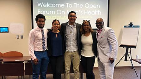 Five individuals standing in front of a screen with a welcome message about a men's health forum.