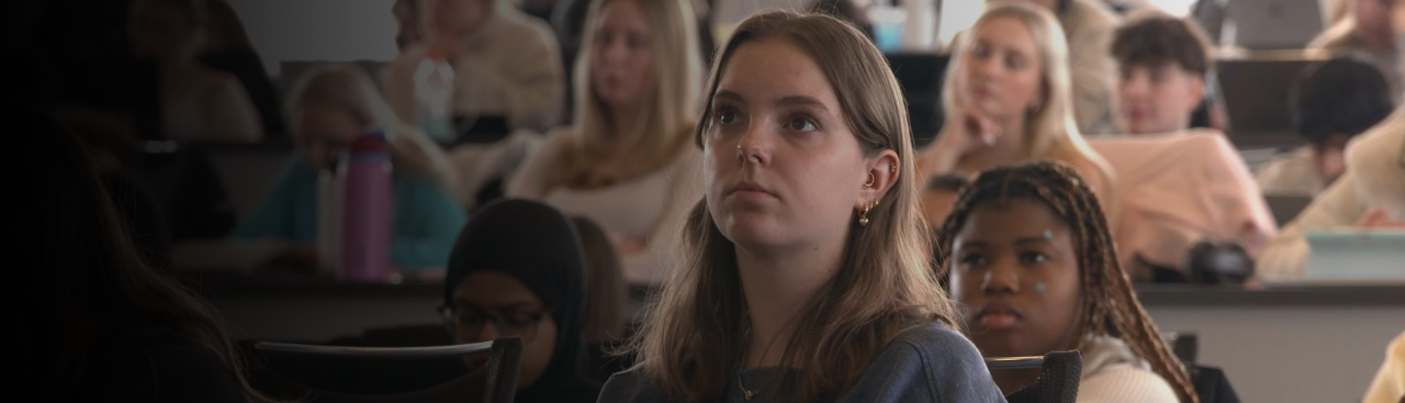 Many students attending a lecture, with one student in focus listening attentively to the instructor.