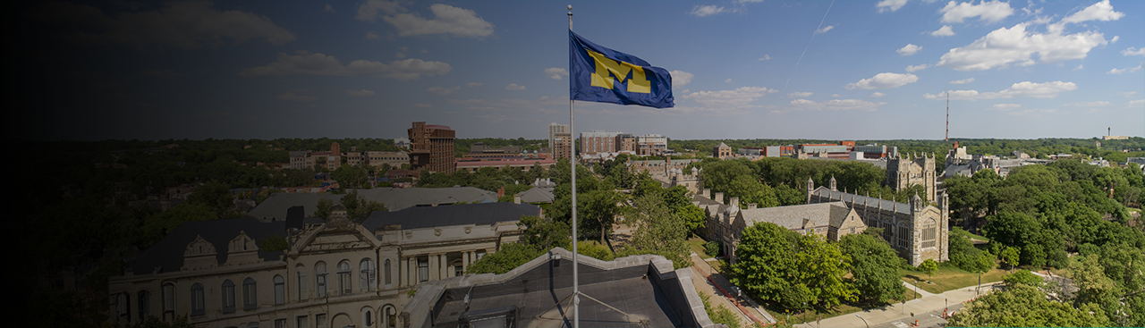 Aerial view of the University of Michigan campus with a UM flag prominently waving in the foreground."