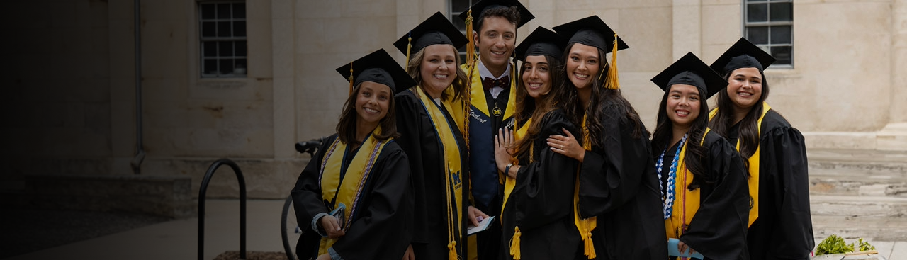 Nursing graduates in cap and gown smiling and posing outside a University of Michigan building.