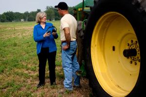 A woman talks to a man outside by a tractor while holding a pair of headphones.