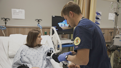 Male nursing student in scrubs works with a patient in a bed. 