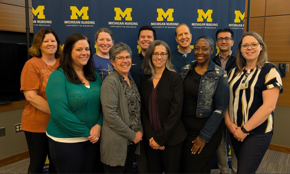 Faculty from both departments smiling together in from of a Michigan Nursing background