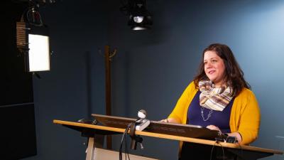 Female instructor stands at a desk in front of a camera 