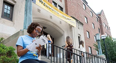 A student stands outside a dorm during Welcome Week, reviewing a packet of information as other students walk past her and up the steps into the building’s entrance.